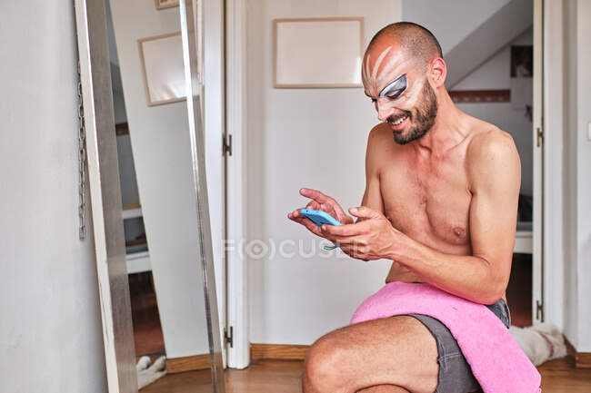 Homme barbu torse nu excentrique avec drag queen maquillage et bracelet de fierté en écrivant un message sur téléphone portable bleu tout en étant assis près du miroir à la maison — Photo de stock