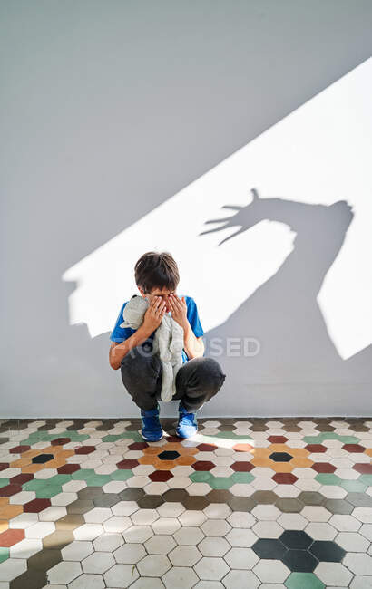 Menino assustado com brinquedo nas mãos sentado perto da parede com sombra de pais violentos com braço levantado — Fotografia de Stock