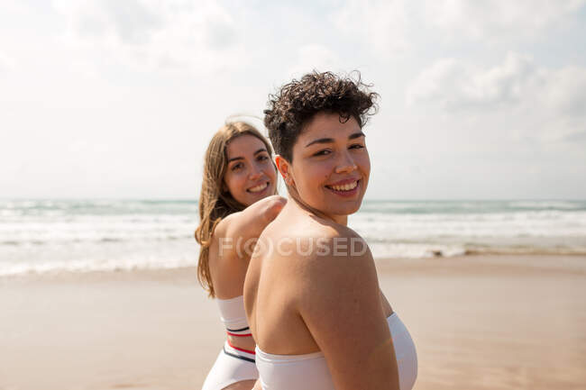 Jovens namoradas felizes em roupa de banho na costa arenosa do mar ondulado sob céu nublado em dia ensolarado olhando para a câmera — Fotografia de Stock