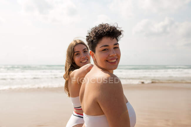 Glückliche junge Freundinnen in Badebekleidung an der sandigen Küste des welligen Meeres unter wolkenverhangenem Himmel an einem sonnigen Tag mit Blick in die Kamera — Stockfoto