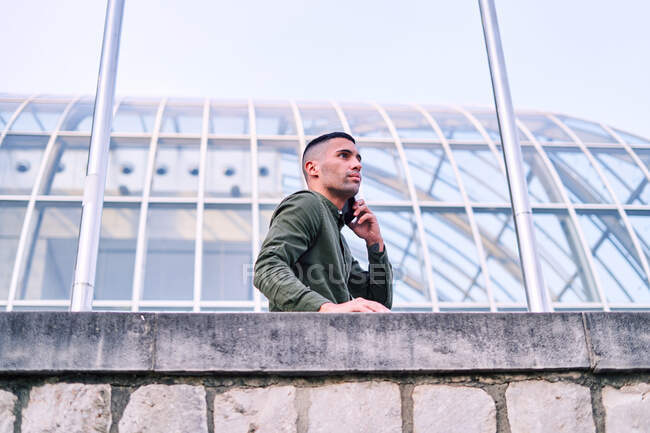 Niedriger Winkel eines erwachsenen hispanischen Mannes im Hemd, der sich an eine Steinmauer lehnt und außerhalb des modernen Glasgebäudes mit dem Handy spricht — Stockfoto