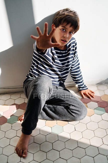 Niño étnico preadolescente infeliz sentado en el suelo y extendiendo el brazo mientras trata de protegerse de los padres enojados - foto de stock