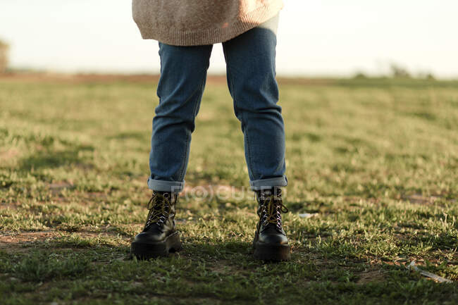 Crop anonymous female traveler wearing knitted sweater with jeans and black leather boots standing in green grassy field in spring countryside — Stock Photo