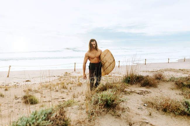 Young surfer man with long hair dressed in wetsuit walking looking away with surfboard in sandy dunes — Stock Photo