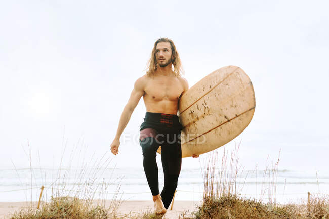 Giovane surfista dai capelli lunghi vestito di muta che cammina distogliendo lo sguardo con tavola da surf in dune sabbiose — Foto stock