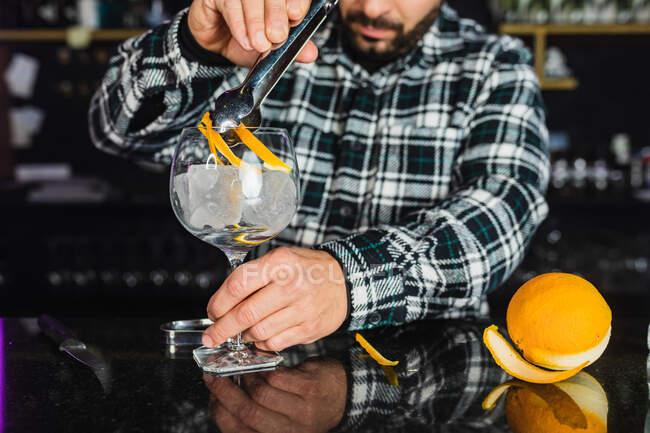 Cropped unrecognizable male barkeeper adding orange peel into a glass while preparing cocktail standing at counter in modern bar — Stock Photo