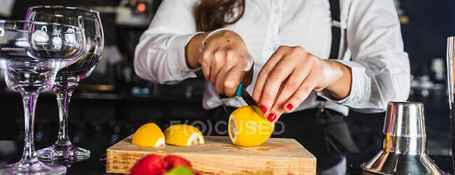 Засеянная неузнаваемая барменша в стильном наряде готовит коктейль с лимонными ломтиками, стоя за стойкой в современном баре — стоковое фото