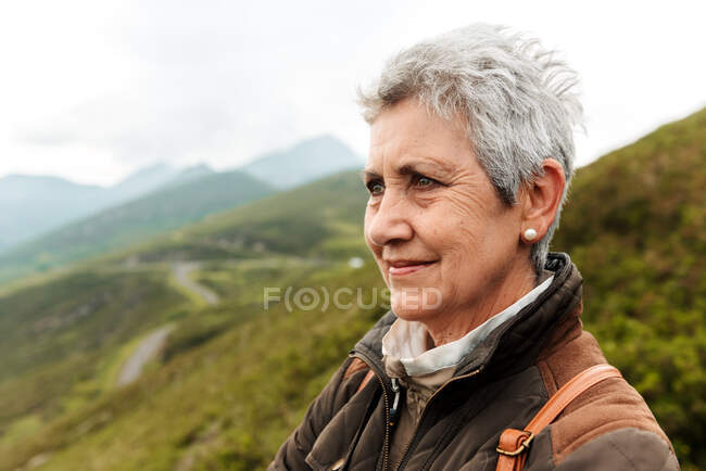 Positive elderly female traveler face and looking away on blurred background of nature — Stock Photo