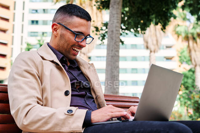 Bajo ángulo de joven hispano enfocado en ropa casual elegante trabajando remotamente en un proyecto independiente en un portátil mientras está sentado con una taza de café cerca de edificios urbanos modernos - foto de stock