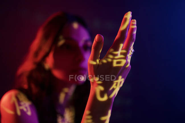 Fashionable young female model with light projection in shape of oriental hieroglyphs looking at extended hand in dark studio with red illumination — Stock Photo