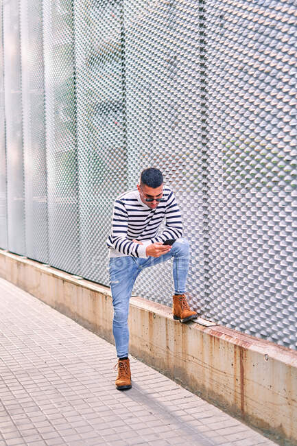 Full body Hispanic man in stylish outfit looking away and using cellphone while leaning on wall on city street — Stock Photo