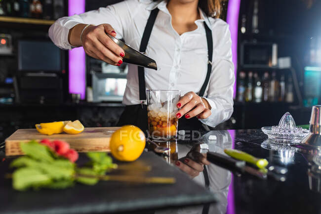 Ritagliato irriconoscibile barista donna in abito elegante aggiungendo cubetti di ghiaccio in vetro mentre prepara cocktail di mojito in piedi al bancone nel bar moderno — Foto stock