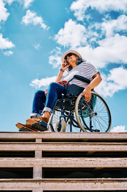 Ältere behinderte Frau sitzt im Rollstuhl und telefoniert in der Nähe der Treppe vor blauem Himmel in der Stadt — Stockfoto