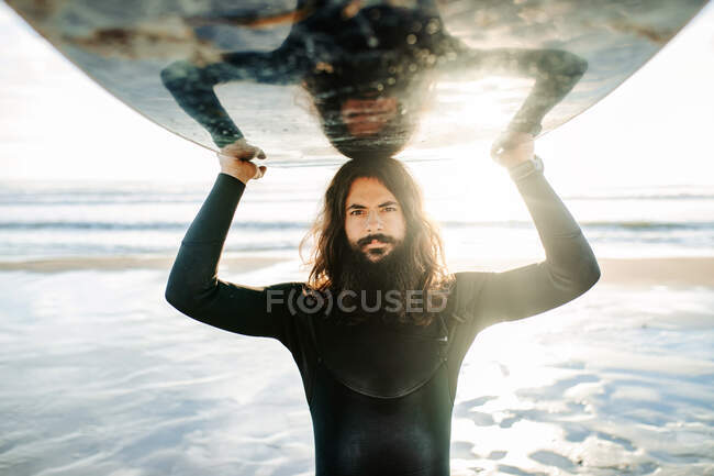 Portrait de jeune surfeur homme aux cheveux longs et barbe vêtu d'une combinaison de plongée debout regardant la caméra sur la plage avec la planche de surf au-dessus de la tête pendant le lever du soleil — Photo de stock