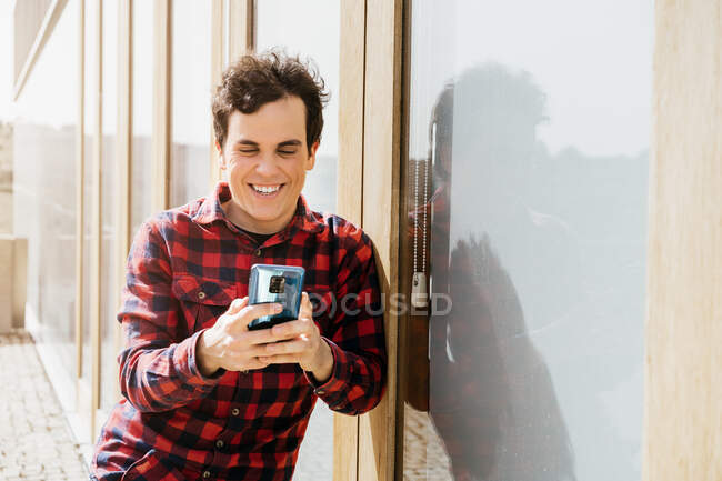 Веселий молодий чоловік у повсякденному носінні використовує смартфон, стоячи проти сучасної міської будівлі — стокове фото