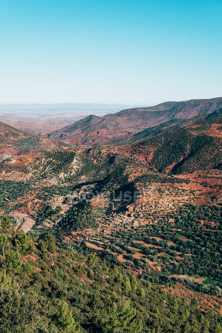 Dall'alto di rosso colorato ricoperto di piante verdi montagne e cielo blu chiaro sullo sfondo in Marocco — Foto stock
