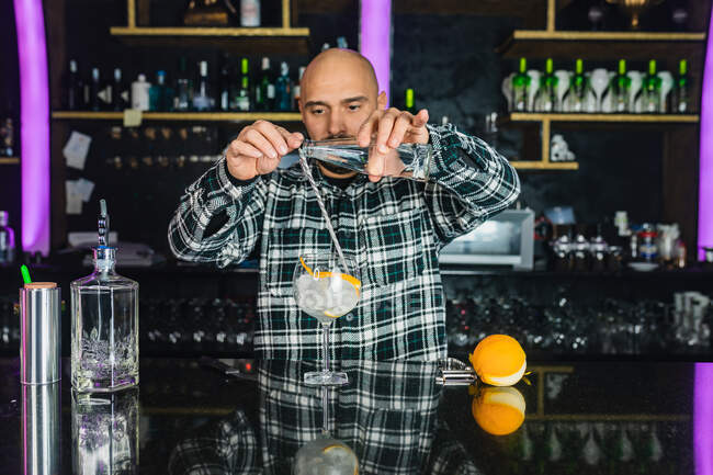 Сосредоточенный бармен-мужчина добавляет жидкость из бутылки в стакан с длинной ложкой во время приготовления коктейля стоя у стойки в современном баре — стоковое фото