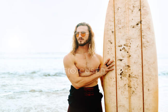 Giovane surfista con i capelli lunghi vestito con muta e occhiali da sole alla moda in piedi guardando lontano con la tavola da surf verso l'acqua per catturare un'onda sulla spiaggia — Foto stock