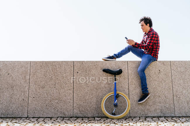 Vista lateral de cuerpo completo del joven con camisa a cuadros y jeans sentados en valla de piedra cerca del monociclo y el teléfono móvil de navegación durante el tiempo libre en la ciudad - foto de stock