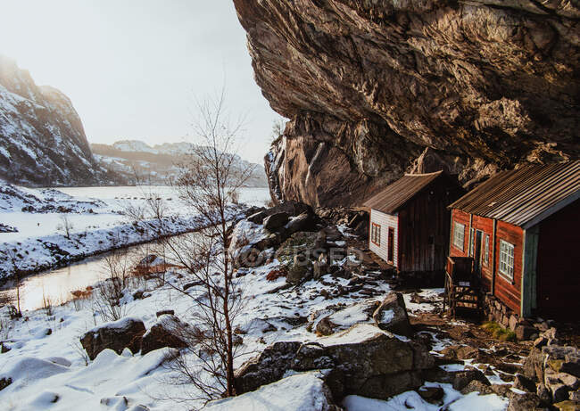 Cabanas envelhecidas perto de paredes de rocha entre terras selvagens na neve perto do rio estreito e céu azul — Fotografia de Stock