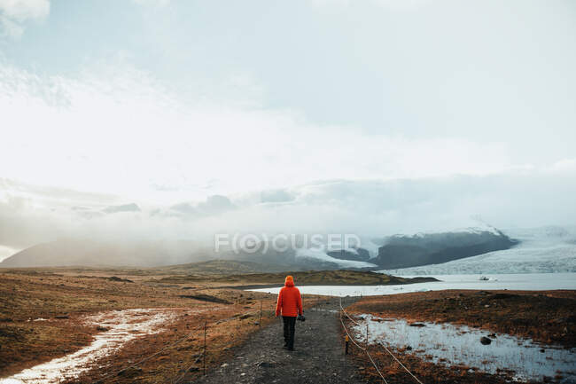 Back view of young tourist going on walkway between wild lands in snow near high stone hills and cloudy sky — Stock Photo