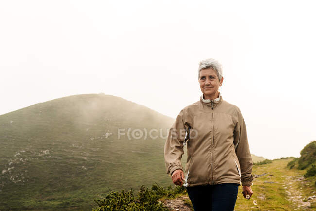Voyageuse âgée avec les cheveux gris courts regardant loin et marchant sur le chemin près de la colline pendant la journée dans la nature — Photo de stock