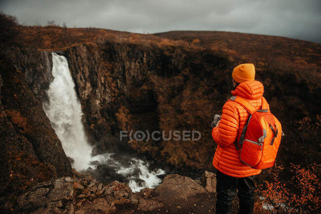 Back view of young tourist in winter wear looking at cascade and mountain river between stone hill — Stock Photo