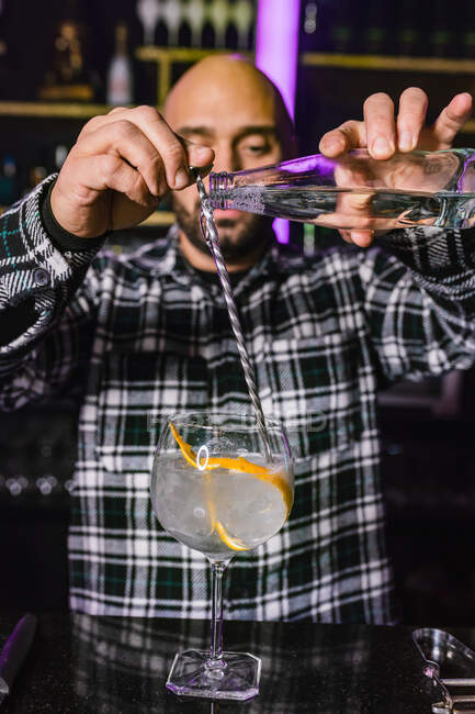 Focused male barkeeper adding liquid from bottle into a glass with long spoon while preparing cocktail standing at counter in modern bar — Stock Photo