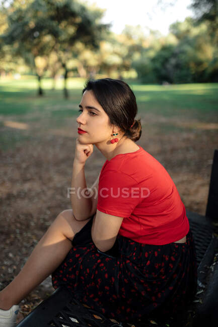 Вид сбоку на очаровательную женщину, сидящую на скамейке и отводящую взгляд, наслаждаясь выходными в летнем парке — стоковое фото