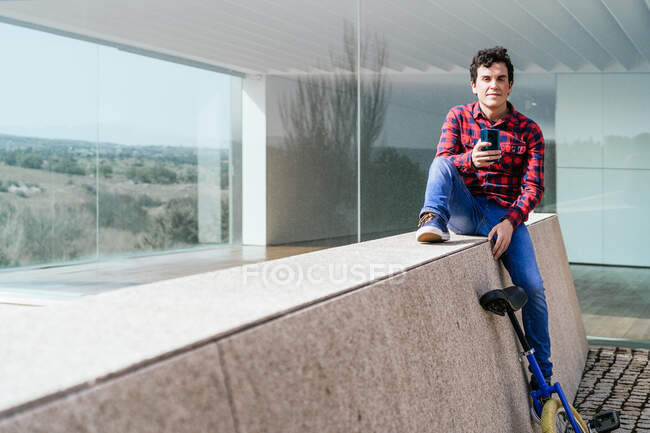 Corpo pieno di giovane ragazzo in camicia a scacchi e jeans seduto sulla recinzione di pietra vicino monociclo e la navigazione cellulare durante il tempo libero in città guardando la fotocamera — Foto stock