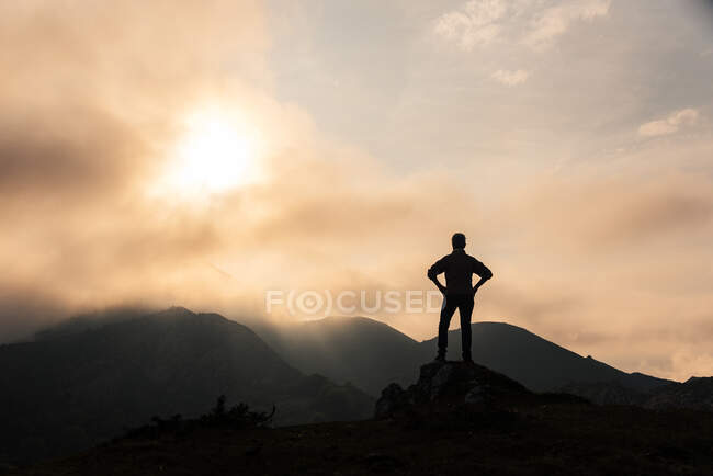 Silhouette eines anonymen Forschers mit den Händen auf der Taille, der bergiges Gelände vor wolkenverhangenem Himmel am Morgen in der Natur bewundert — Stockfoto
