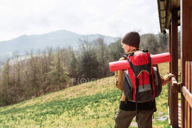 Visão traseira do mochileiro feminino em pé perto da casa de madeira nas montanhas no dia ensolarado — Fotografia de Stock