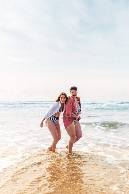 Full body of cheerful girlfriends with mouth opened hugging while standing looking at camera on sandy beach washed by waving sea — Stock Photo