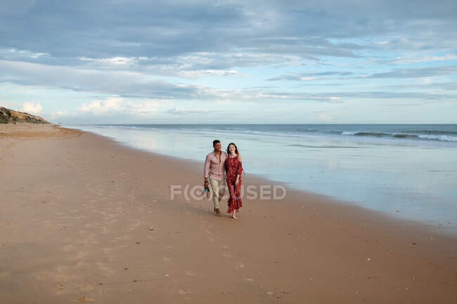 Весела багаторасова пара тримає руки і ходить уздовж мокрого берега, милуючись морем на світанку влітку — стокове фото