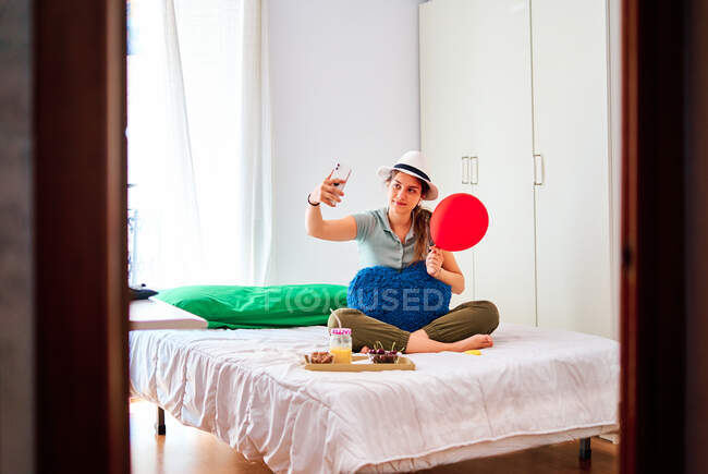 Comprimento total de jovem fêmea em roupas casuais e chapéu segurando balão vermelho e almofada em forma de coração e tomando selfie no smartphone enquanto celebra aniversário sozinho na cama com alimentos e suco — Fotografia de Stock