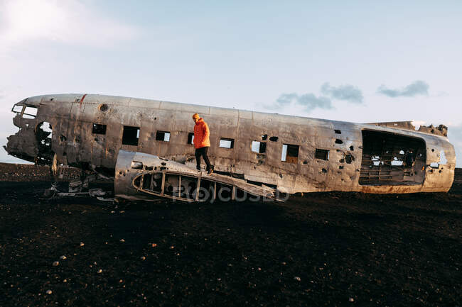 Вид сбоку на молодого туриста, стоящего на разбитом самолете между пустынными землями и голубым небом — стоковое фото