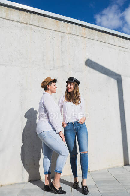 Corpo pieno di giovani amiche positive in abiti e cappelli alla moda che si guardano l'un l'altro sulla passerella vicino al muro grigio nella giornata di sole sotto il cielo blu — Foto stock