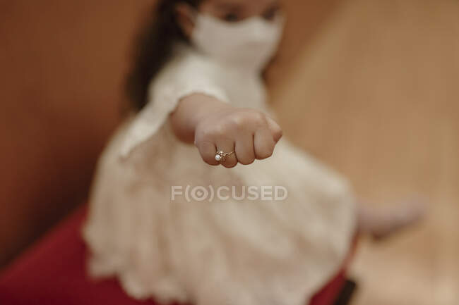 Alto ángulo suave foco de niña encantadora en vestido blanco y máscara protectora mano extendida con anillo elegante con perla en el dedo - foto de stock