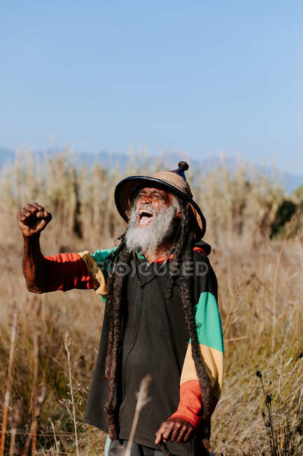 Fröhlicher alter Rastafari mit Dreadlocks mit geschlossenen Augen und geschlossener Faust feiert den Sieg, während er auf einer trockenen Wiese in der Natur steht — Stockfoto