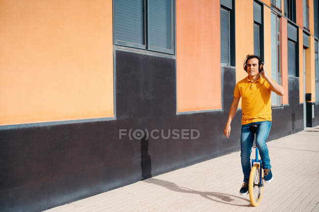 Cuerpo completo de hombre joven moderno con camisa naranja brillante y jeans escuchando música a través de auriculares inalámbricos mientras monta monociclo en el pavimento cerca del edificio urbano - foto de stock