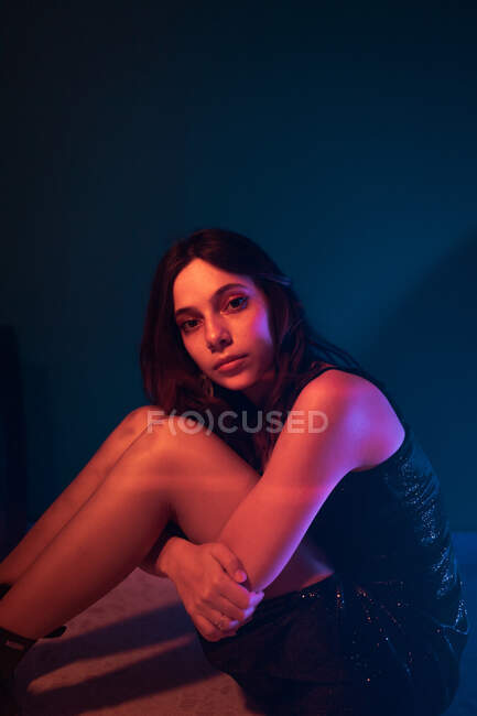 Vista lateral de tranquilo jovem modelo feminino no vestido sentado no chão olhando para a câmera no estúdio escuro com luzes coloridas — Fotografia de Stock