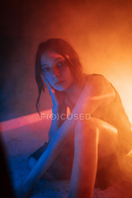 Спокойная молодая модель в платье сидит на полу и опирается на руку, глядя на камеру в темной студии с красочными огнями — стоковое фото
