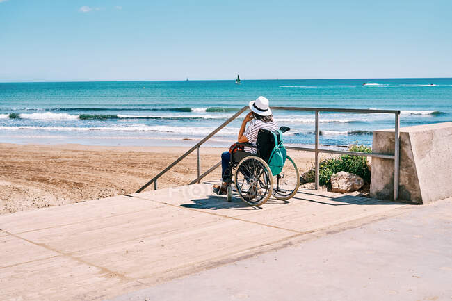 Вид збоку жінка мандрівник в інвалідному візку з рюкзаком насолоджується літньою подорожжю на пляжі біля блакитного моря — стокове фото
