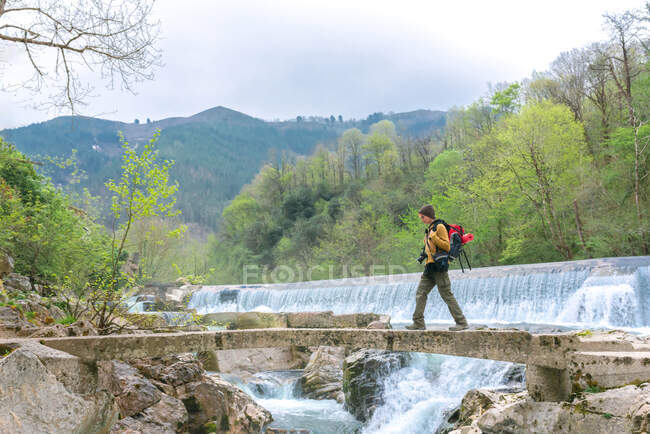 Vista lateral do aventureiro com mochila andando ao longo da ponte de pedra perto de cachoeira em terreno montanhoso — Fotografia de Stock