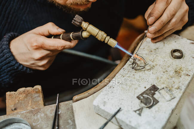 Анонимный ювелир, использующий паяльную лампу для нагрева крошечного металлического орнамента при изготовлении ювелирных изделий на верстаке — стоковое фото