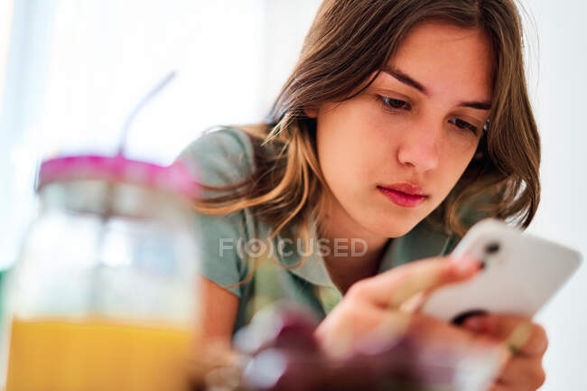 Молода студентка переглядає соціальні мережі на мобільному телефоні біля столу зі свіжими фруктами та соком, проводячи ранок вдома — стокове фото