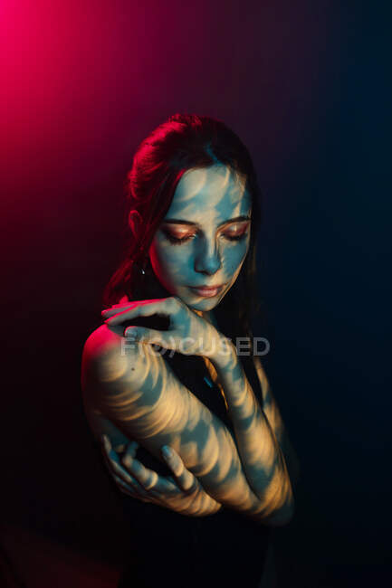 Fashionable young female model with light projection in shape of oriental hieroglyphs looking down in dark studio with red illumination — Stock Photo