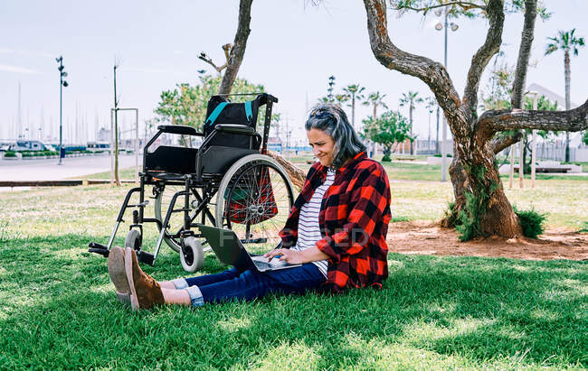 Full body of positive gray haired female with laptop on knees sitting on green grass near wheelchair in park — Stock Photo