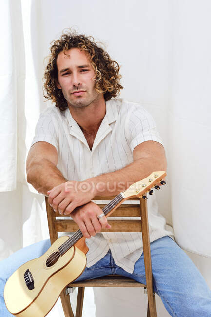 Hombre sereno con el pelo rizado sentado en la silla en la pared blanca con ukelele mirando a la cámara - foto de stock