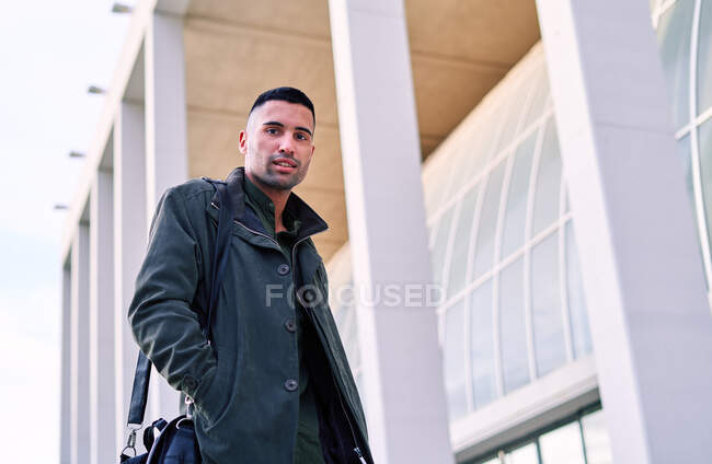 Desde abajo empleado masculino hispano con maletín sonriendo y mirando a la cámara mientras camina sobre el pavimento cerca de un edificio moderno en la ciudad - foto de stock